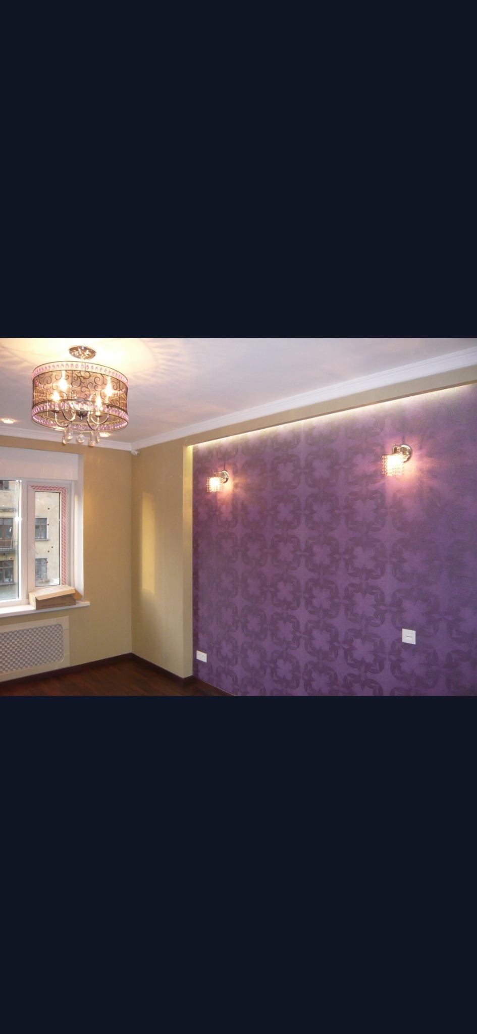 Pokój po odświeżeniu: Fioletowa ściana z wzorem, oświetlona kinkietami i listwą LED, kontrastuje z beżową ścianą przy oknie z grzejnikiem.