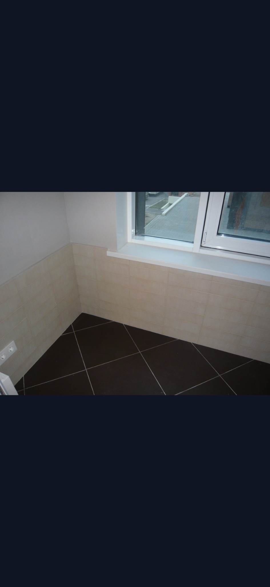 Wykończona łazienka z beżowymi płytkami ściennymi do połowy wysokości i ciemnobrązową podłogą z jasnymi fugami ułożoną w romby, widok na okno z białymi ramami.