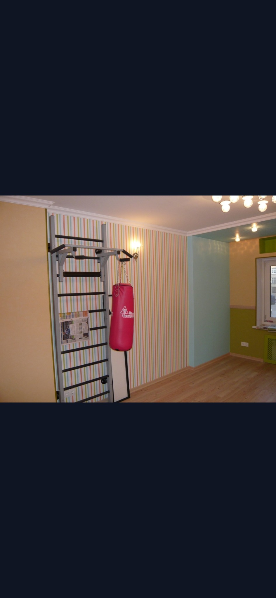 Pokój z elementami sportowymi: drabinka gimnastyczna z gazetą i różowy worek bokserski wiszące na ścianie w pionowe paski, obok ściana żółta i fragment ściany pomalowany na zielono, drewniana...