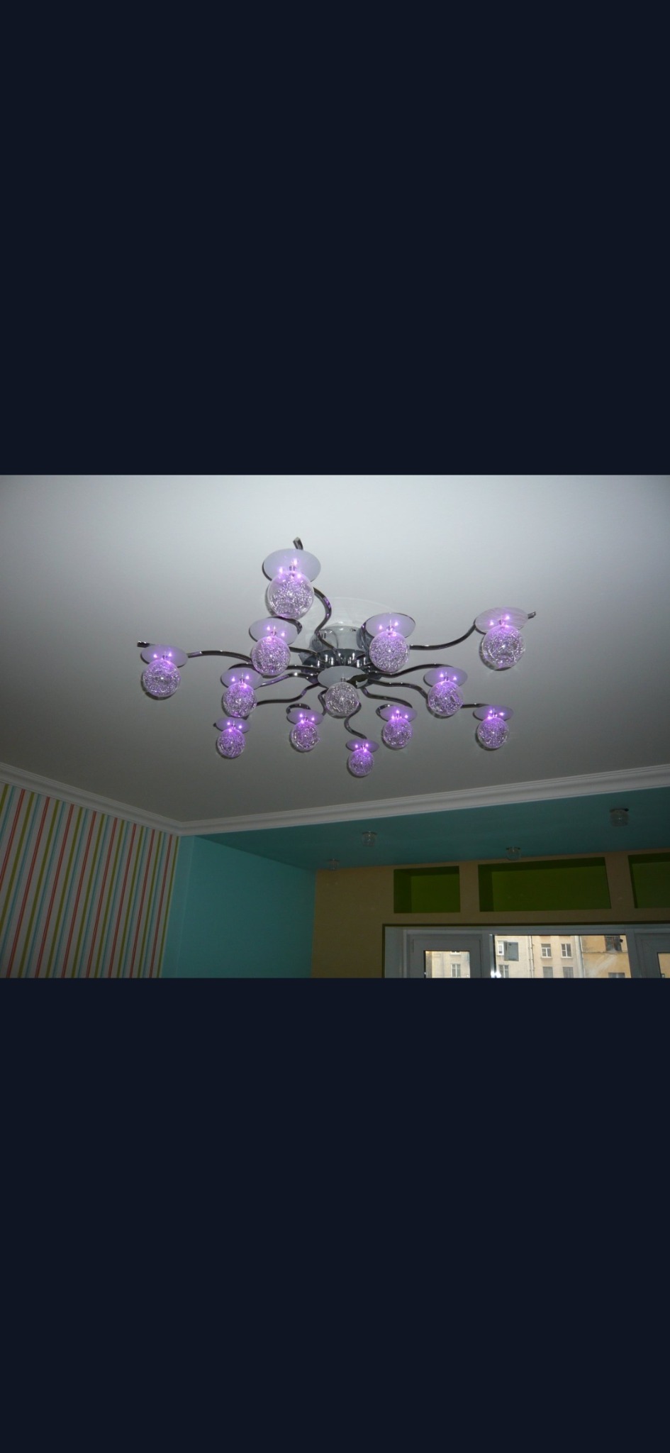 Sufitowa lampa z wieloma szklanymi kloszami oświetlonymi fioletowym światłem, widoczna na tle pomalowanego pokoju z kolorowymi ścianami.