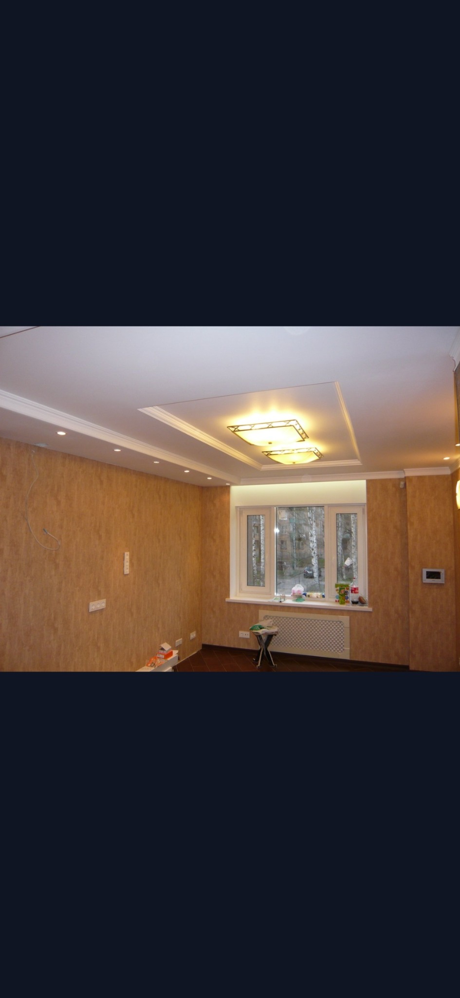 Pokój po remoncie z wykończonym sufitem podwieszanym z oświetleniem, ścianami pokrytymi tapetą o fakturze drewna i nową podłogą, widoczne puszki elektryczne i grzejnik pod oknem.