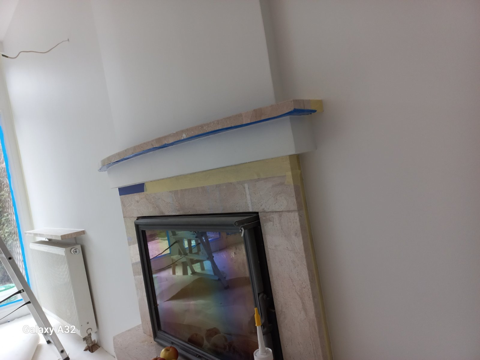 Fragment ściany z kominkiem w trakcie obkładania kamieniem, zabezpieczonym taśmą malarską, widoczny grzejnik i drabina.