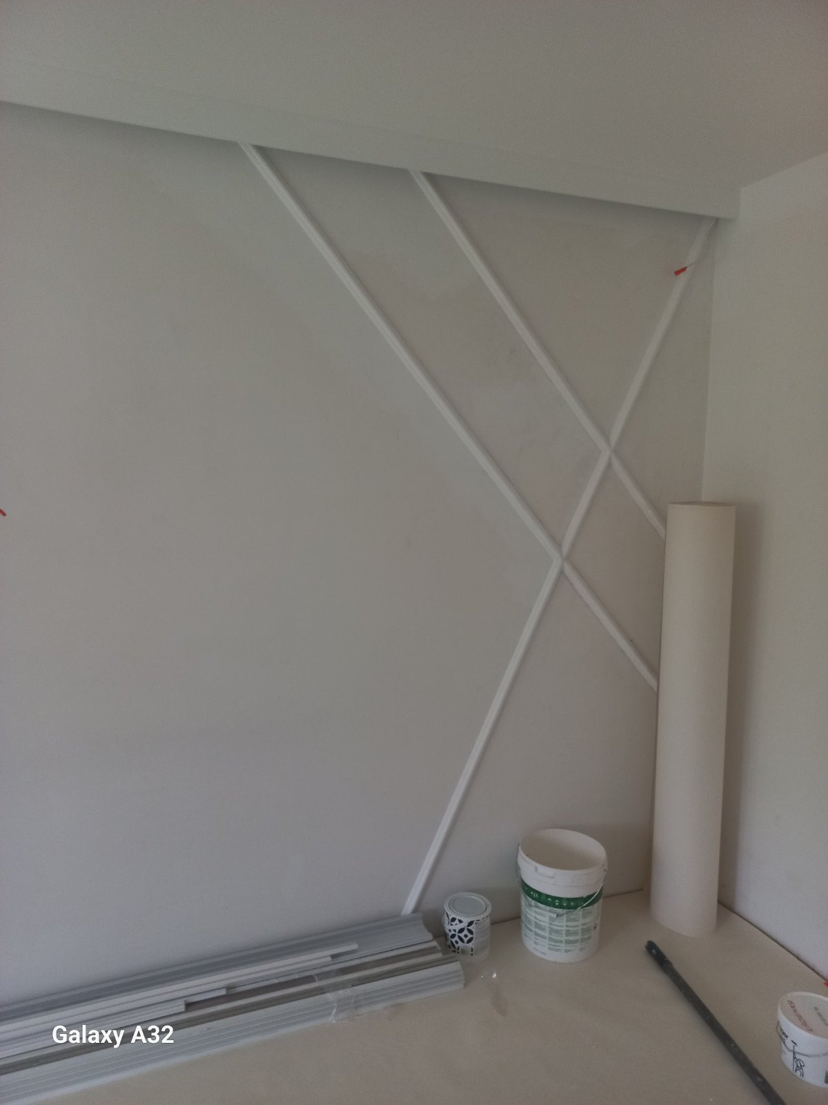 Ściana z dekoracyjnymi listwami w geometryczny wzór, widoczne puszki z farbą, wałek malarski i listwy przypodłogowe przed montażem.