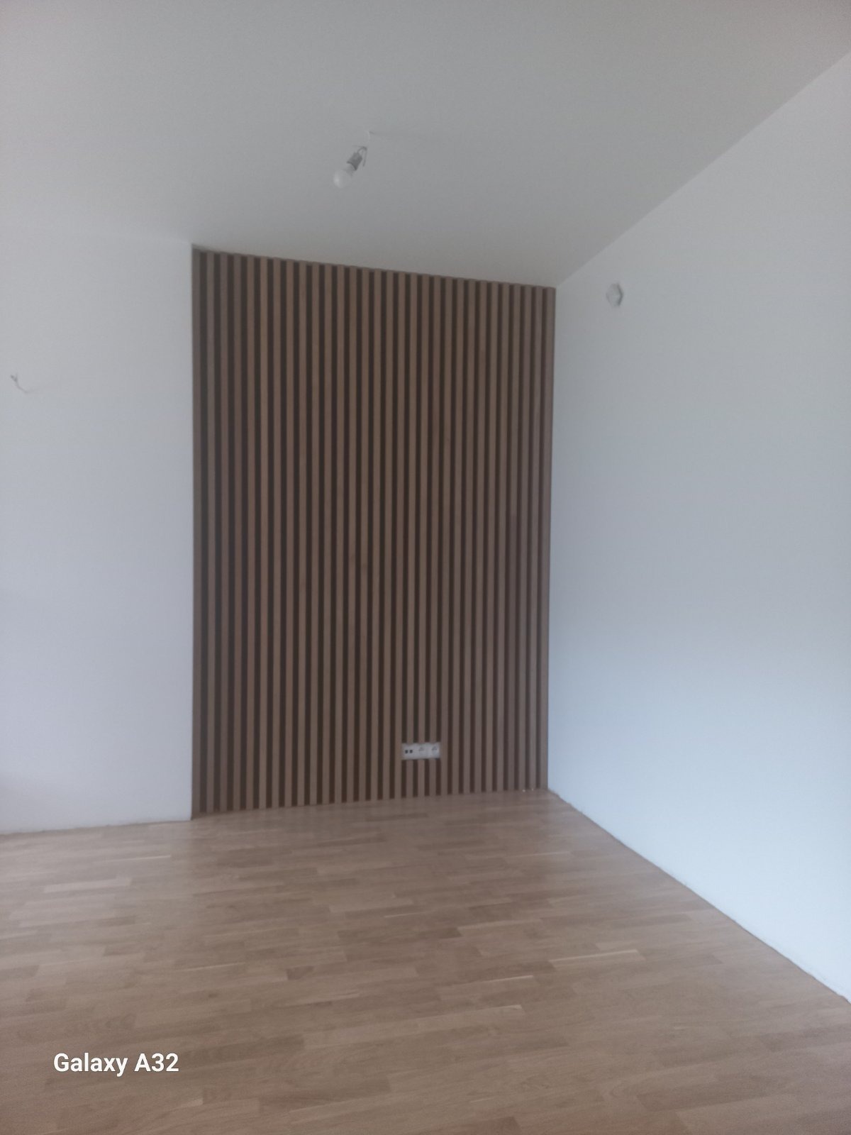 Ściana wykończona panelami dekoracyjnymi z pionowych listew w odcieniach brązu, na podłodze parkiet, białe ściany i widoczny kontakt elektryczny.