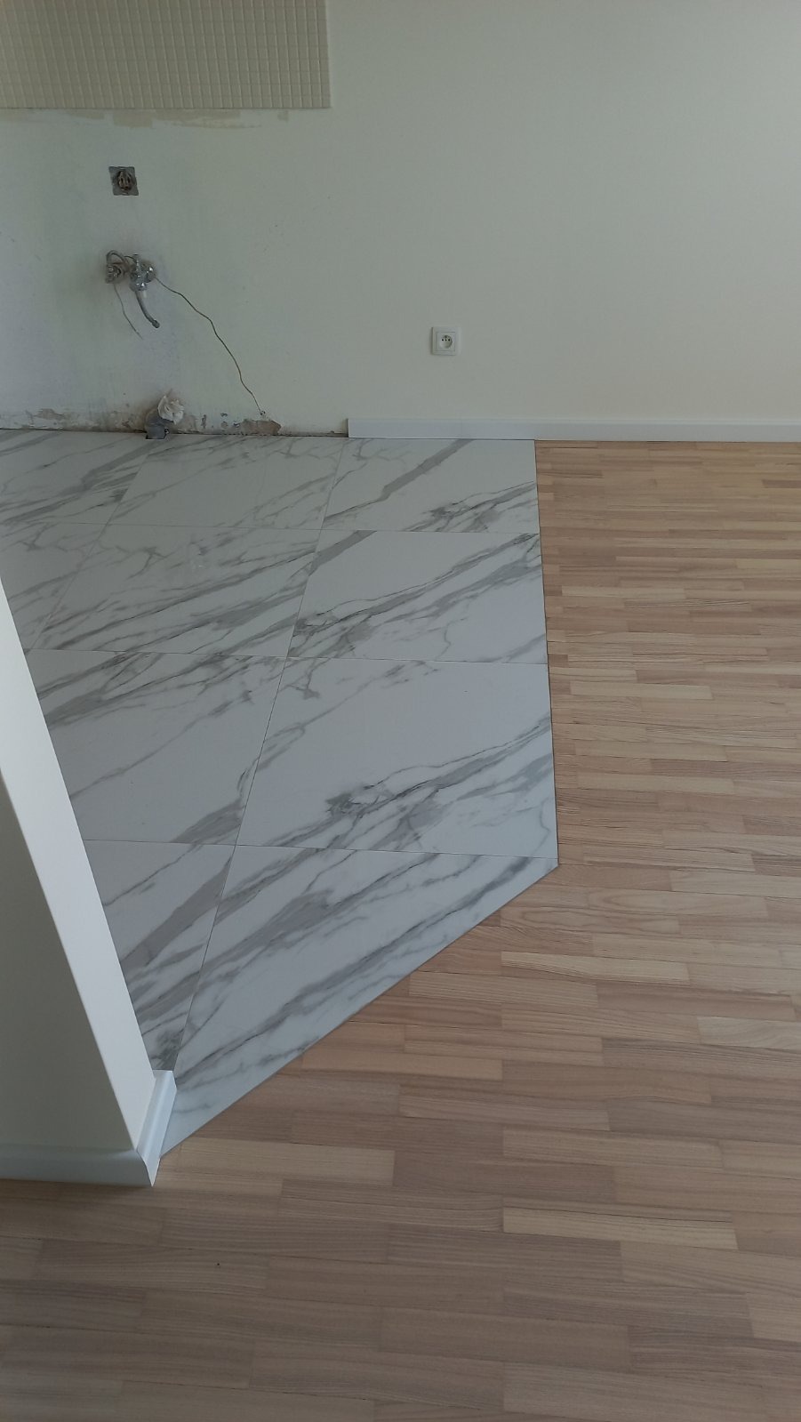 Połączenie dwóch rodzajów podłóg: marmurowe płytki i drewnopodobne panele, widoczne niedokończone prace przy ścianie z wystającymi przewodami.