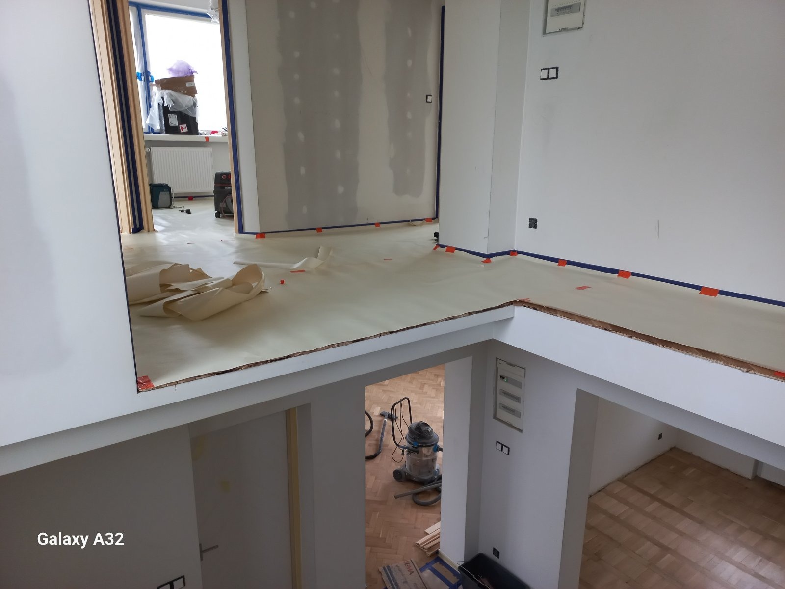 Wnętrze w trakcie prac przygotowawczych: zabezpieczona podłoga folią i taśmą, ściany przygotowane do malowania, widoczny odkurzacz przemysłowy i resztki materiałów budowlanych w pomieszczeniu...