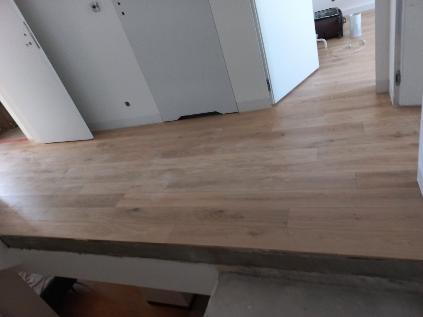 Nowo położona podłoga z paneli w jasnym odcieniu drewna, widoczne białe drzwi i fragment schodów z szarą krawędzią.