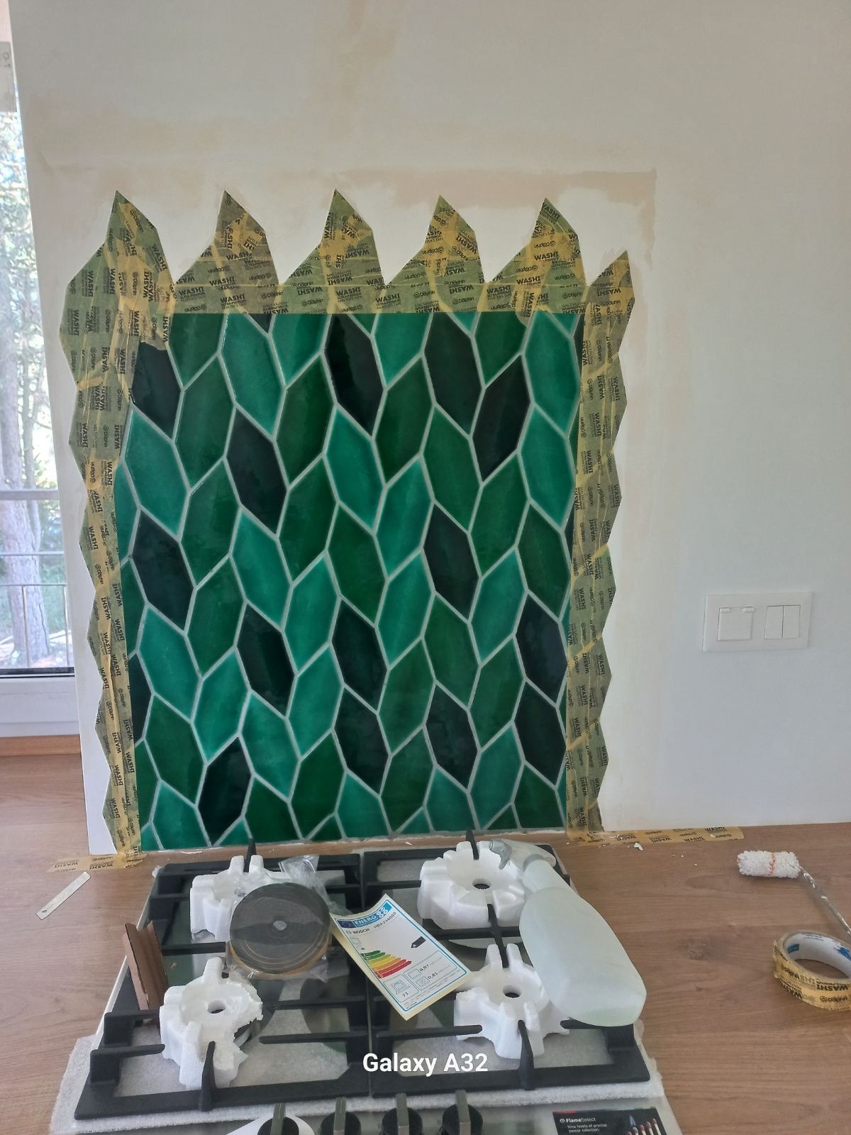 Układanie zielonych, romboidalnych płytek na ścianie, zabezpieczonych taśmą malarską, widoczny fragment kuchenki gazowej i akcesoria glazurnicze.