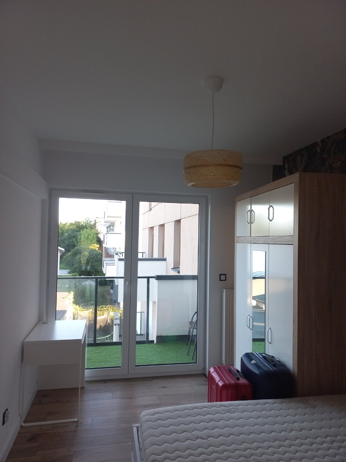 Jasne wnętrze pokoju z wyjściem na balkon z sztuczną trawą, widoczna szafa, biurko i lampa z abażurem z naturalnego materiału.