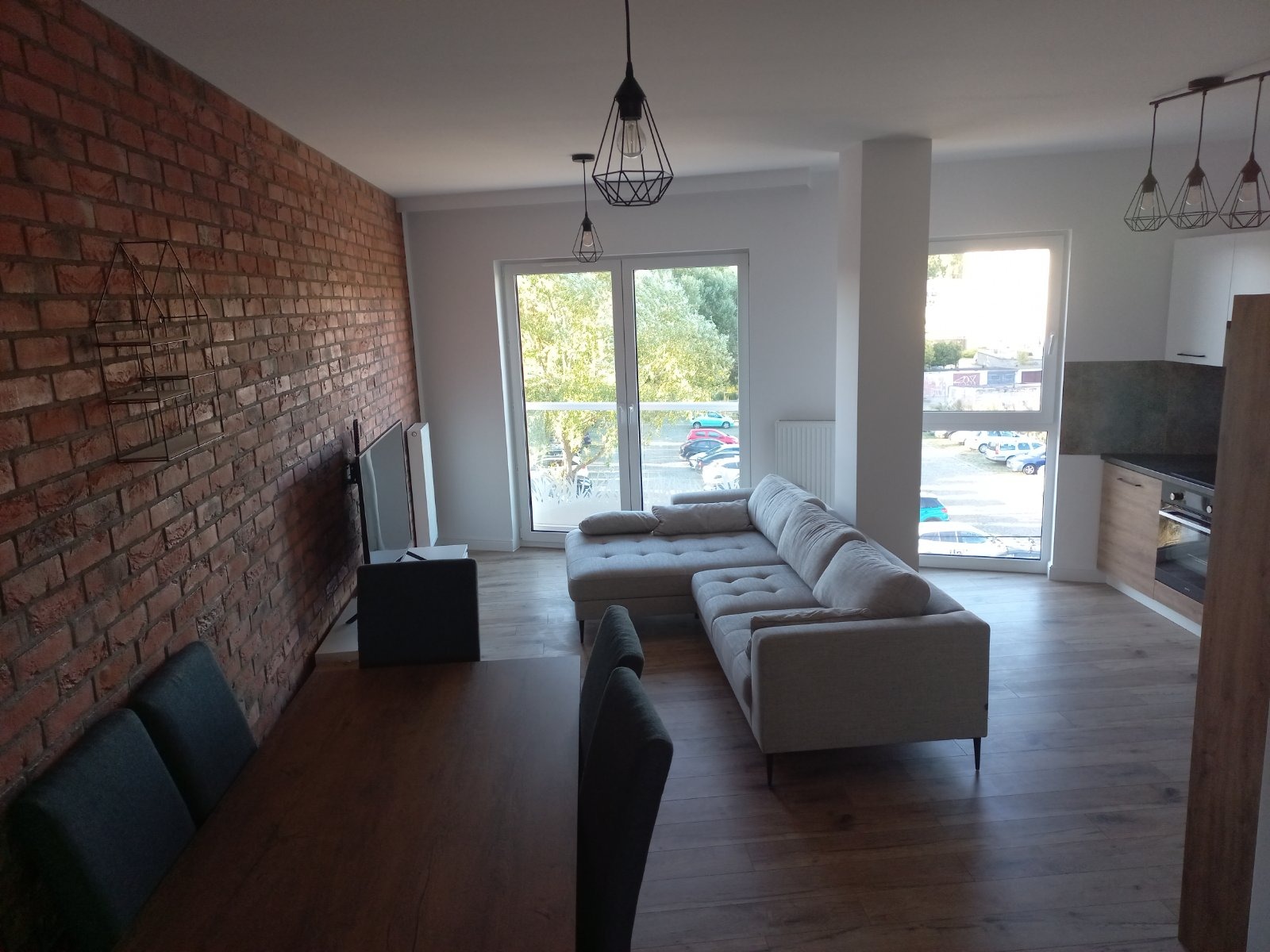 Nowoczesny salon z ceglaną ścianą, szarą sofą narożną, drewnianym stołem i designerskimi lampami wiszącymi, widok na zewnątrz przez duże okna.