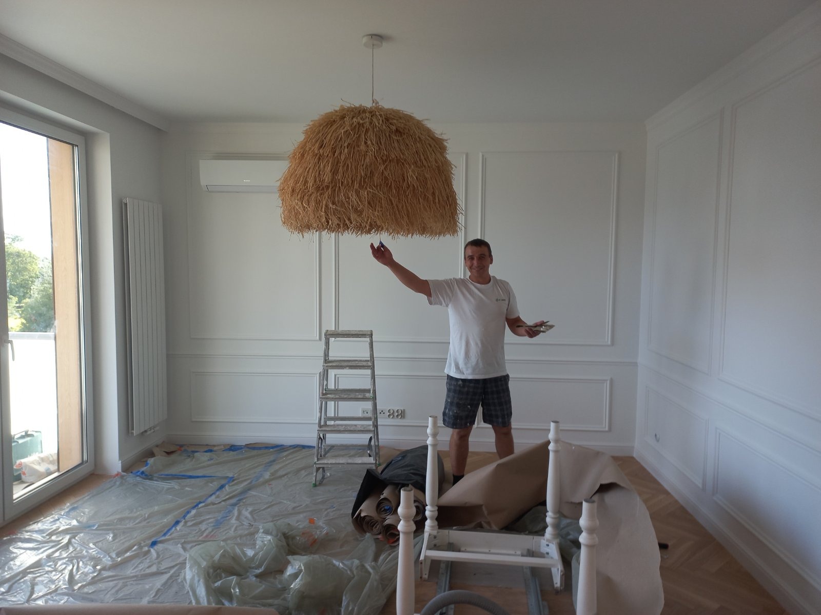 Malarz z pędzlem i paletą w dłoni, stojący w pokoju z nowo pomalowanymi białymi ścianami z dekoracyjnymi listwami, pod słomkową lampą, z podłogą zabezpieczoną folią malarską.