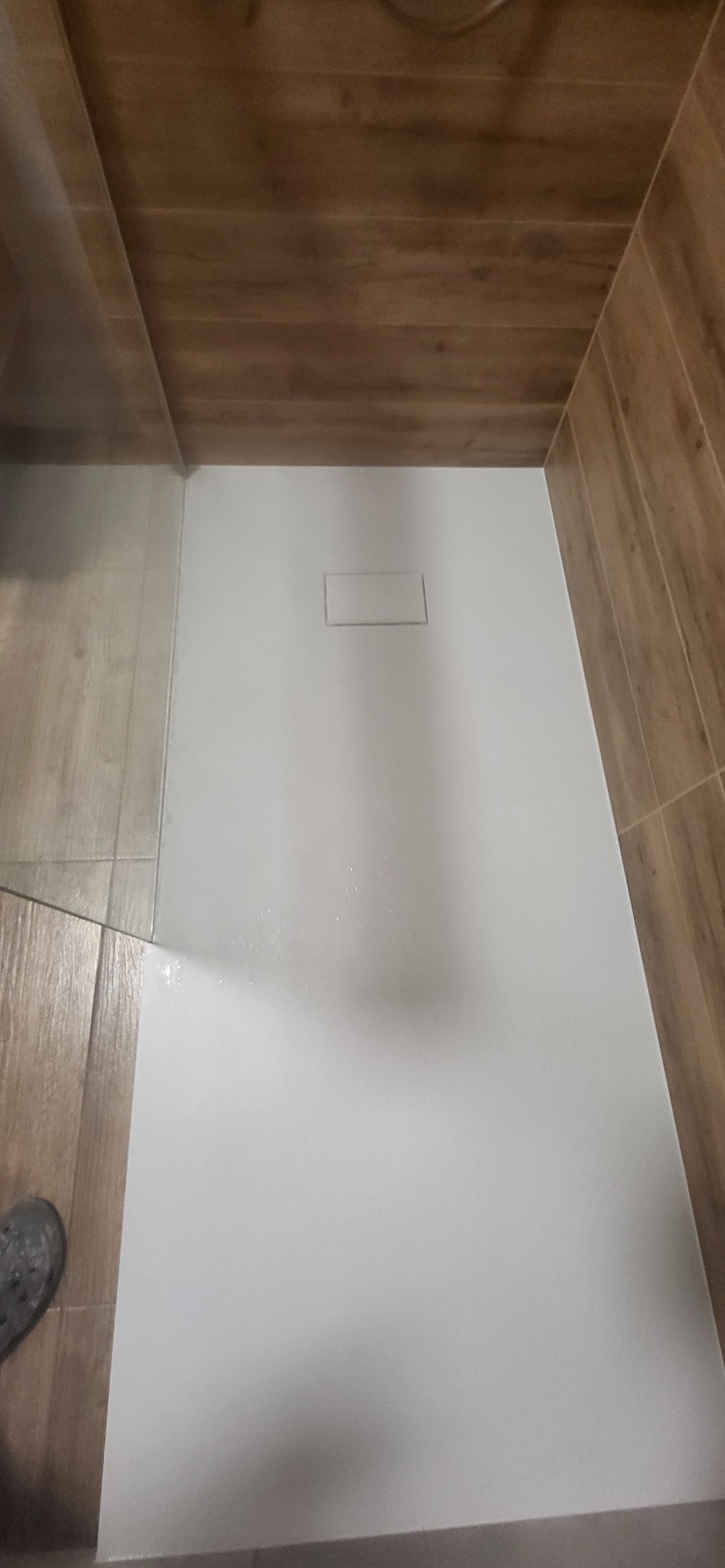 Nowoczesny biały brodzik z odpływem liniowym, otoczony brązowymi płytkami imitującymi drewno, widoczne niedoskonałości na powierzchni brodzika.