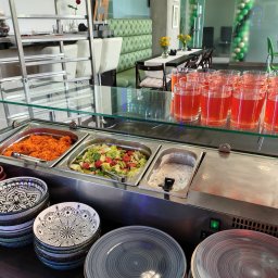 Bistro Cybernetyki - Bufet samoobsługowy z sałatkami, surówką z marchewki i sosem, uzupełniony o stosy dekoracyjnych talerzy i rzędy napojów w szklankach, w tle widoczne stoliki restauracyjne.