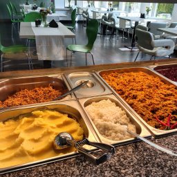 Bistro Cybernetyki - Bufet z różnorodnymi potrawami: puree ziemniaczane ze specjalną łyżką do nakładania, ryż, kasza gryczana, bigos i danie mięsne z papryczkami chili, w tle widoczne stoliki i krzesła w jasnej sali.