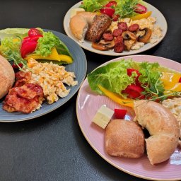 Bistro Cybernetyki - Trzy talerze z różnorodnymi śniadaniami: bajgle, jajecznica, bekon, kiełbasa, pieczarki, ser, sałata i warzywa, podane na talerzach w kolorach szarym, białym i różowym na czarnym tle.