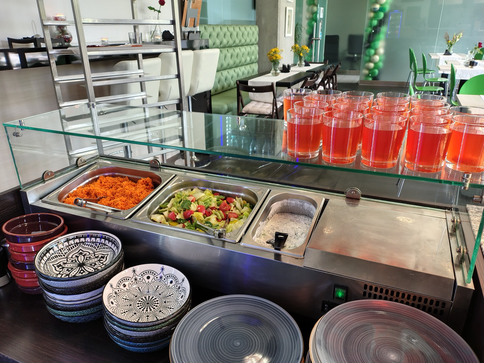 Bufet samoobsługowy z sałatkami, surówką z marchewki i sosem, uzupełniony o stosy dekoracyjnych talerzy i rzędy napojów w szklankach, w tle widoczne stoliki restauracyjne.