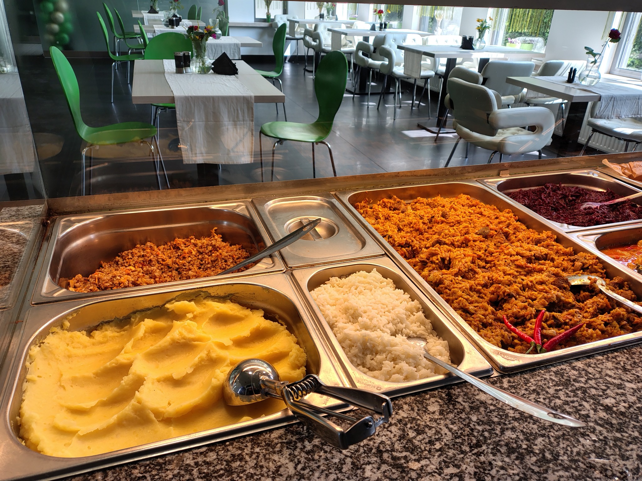 Bufet z różnorodnymi potrawami: puree ziemniaczane ze specjalną łyżką do nakładania, ryż, kasza gryczana, bigos i danie mięsne z papryczkami chili, w tle widoczne stoliki i krzesła w jasnej sali.