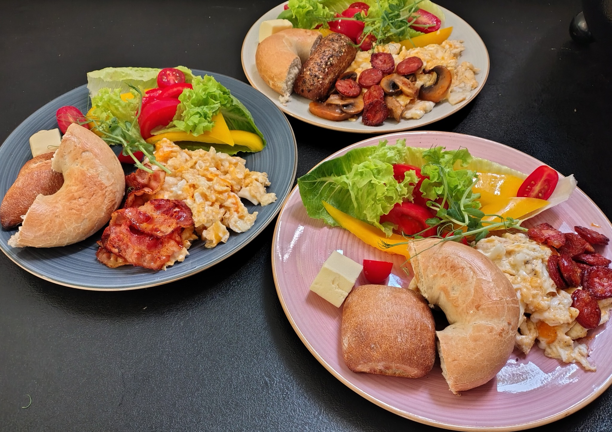 Trzy talerze z różnorodnymi śniadaniami: bajgle, jajecznica, bekon, kiełbasa, pieczarki, ser, sałata i warzywa, podane na talerzach w kolorach szarym, białym i różowym na czarnym tle.