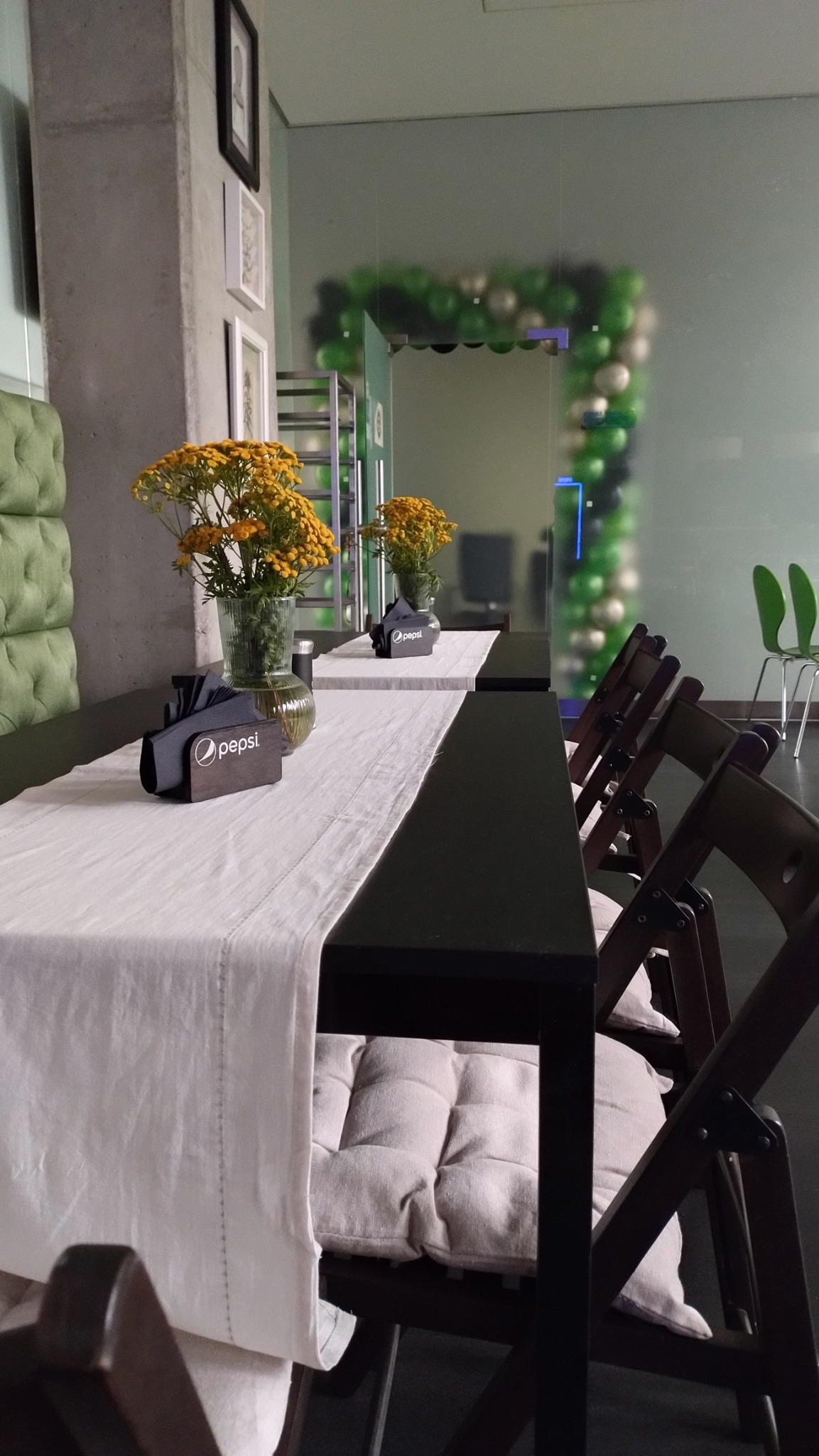 Uroczystość w lokalu: Długi stół z czarnym blatem, nakryty białym obrusem, ozdobiony wazonami z żółtymi kwiatami i serwetnikami z logo Pepsi, krzesła z poduszkami, w tle dekoracja z zielonych...