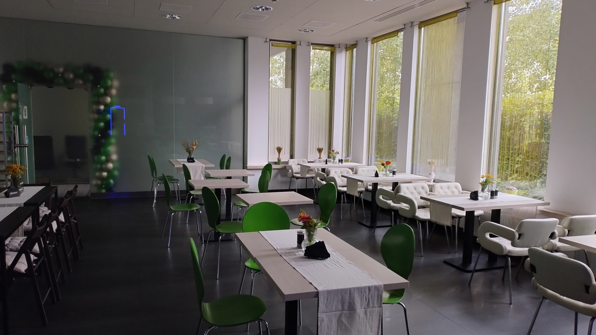 Jasne wnętrze sali z ustawionymi stołami i krzesłami, dekoracjami kwiatowymi na stołach, widok na zieleń za oknami, w tle łuk z balonów.