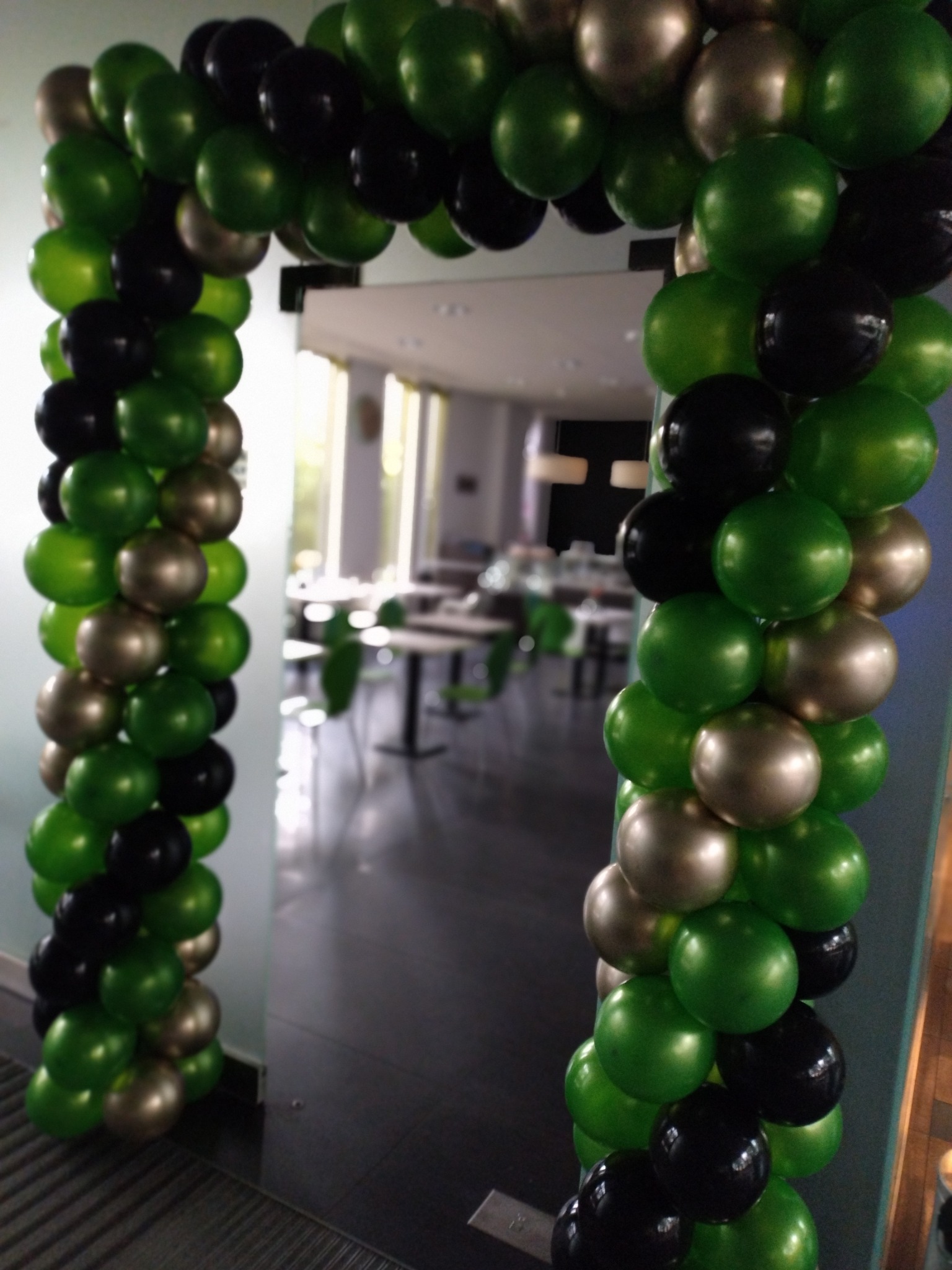 Dekoracja wejścia do sali balonami w kolorach zielonym, czarnym i złotym, widok z bliska, w tle stoliki i krzesła.
