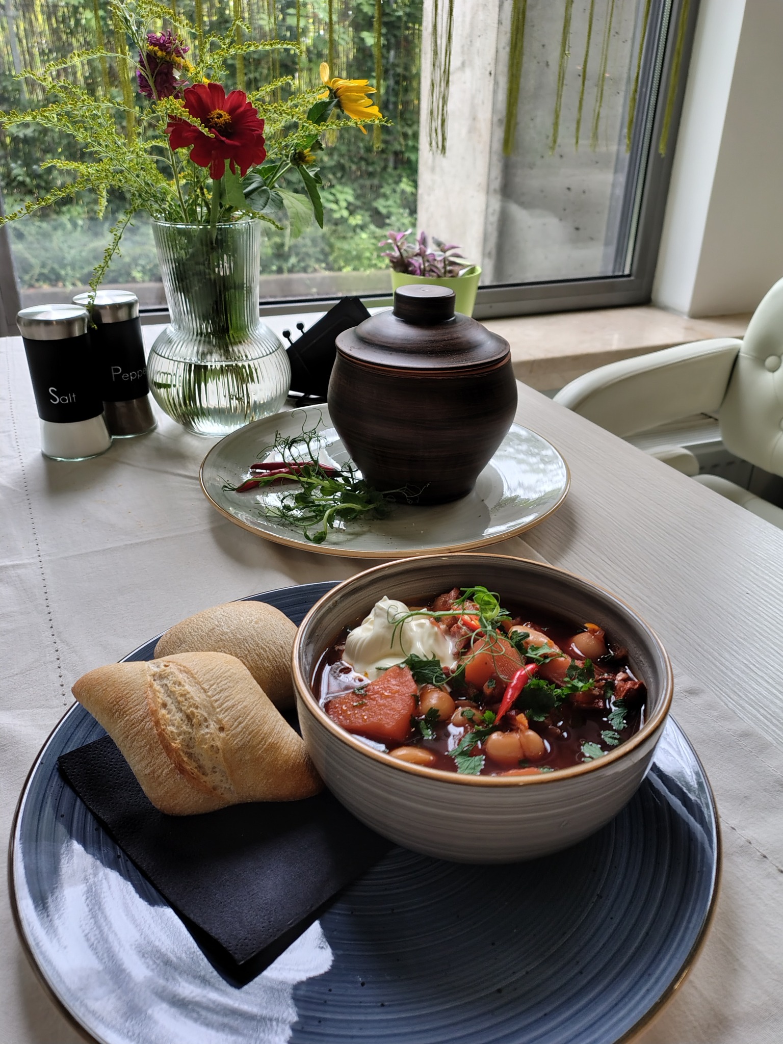Zupa gulaszowa z fasolą, papryką i śmietaną w ceramicznej misce, podana z dwoma bułkami na czarnej serwetce i talerzu, obok ceramiczny garnek, w tle wazon z kwiatami i okno.