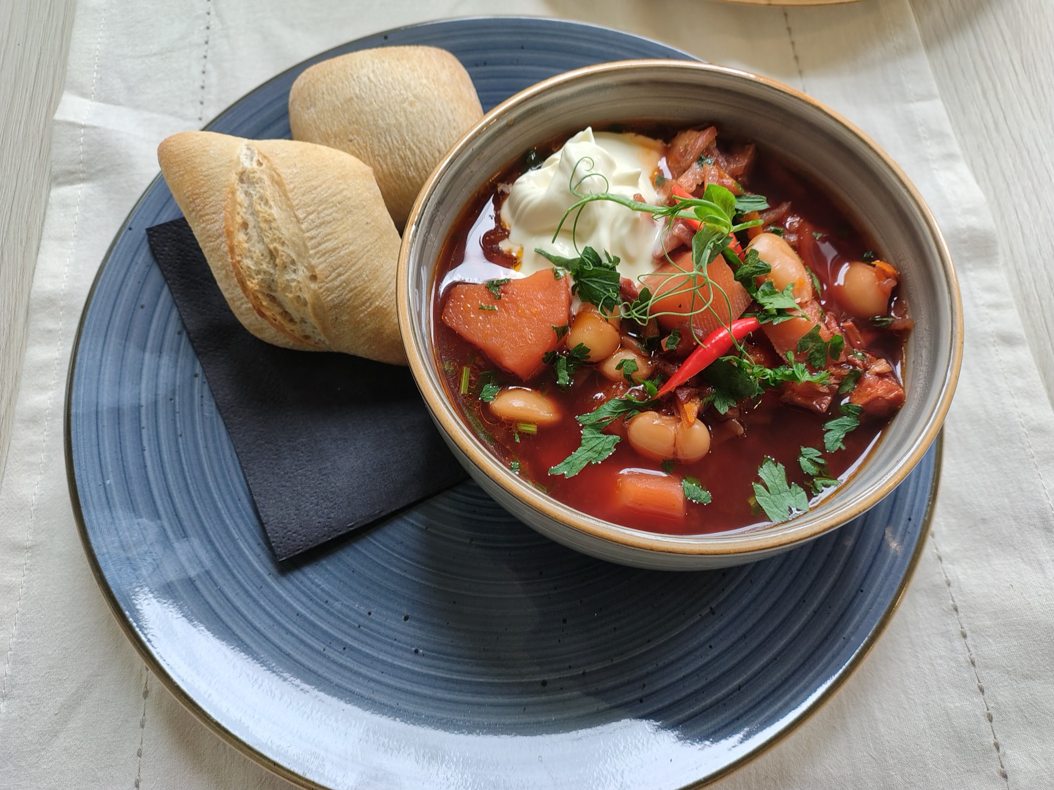 Bordowa zupa z fasolą, warzywami i kleksem śmietany w ceramicznej misce, podana na niebieskim talerzu z dwoma bułeczkami i czarną serwetką na białym obrusie.
