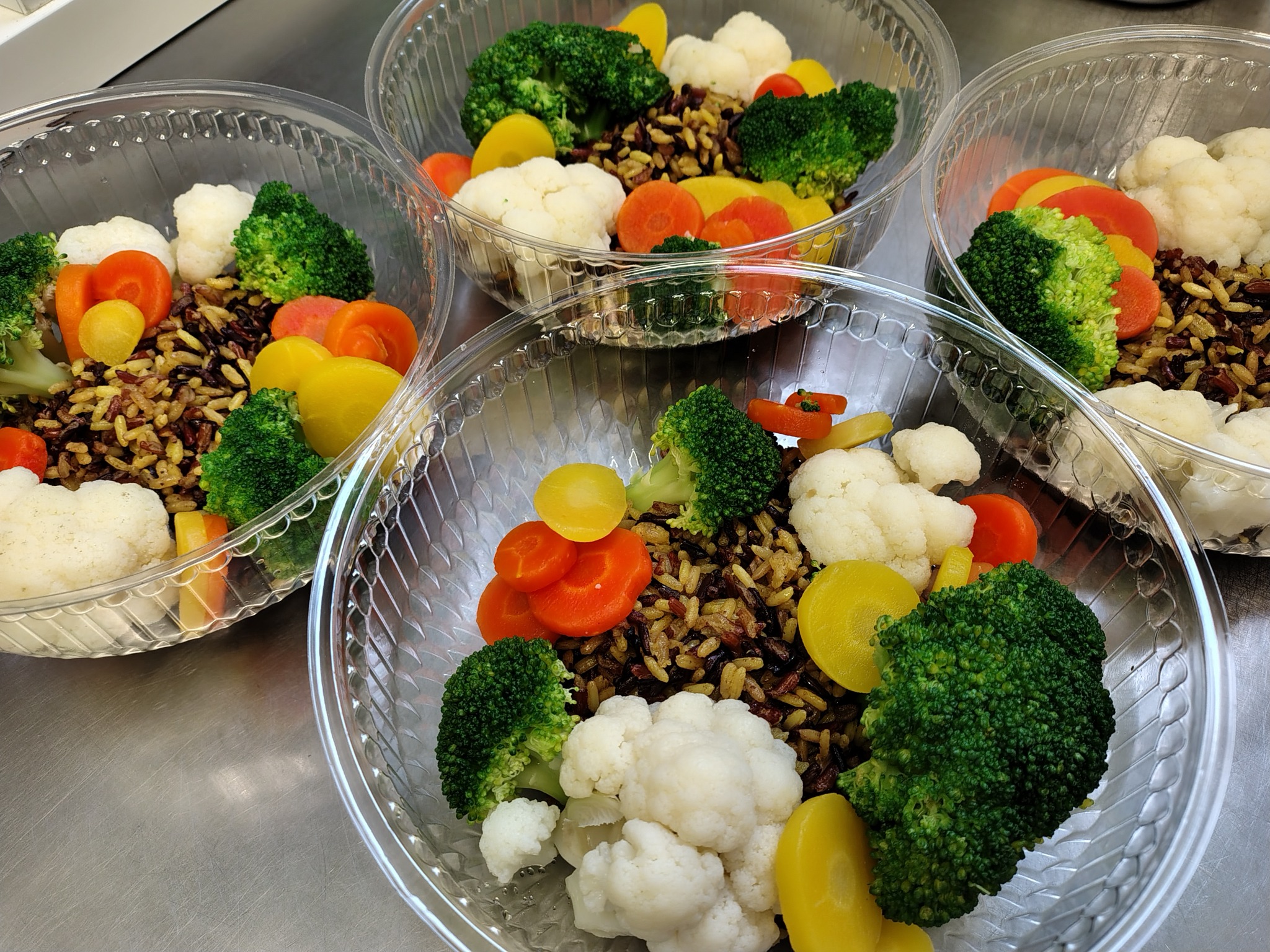 Cztery przezroczyste miski z gotowymi porcjami sałatki z dzikiego ryżu, różyczek brokuła i kalafiora oraz plasterków marchewki i cukinii na metalowym blacie.