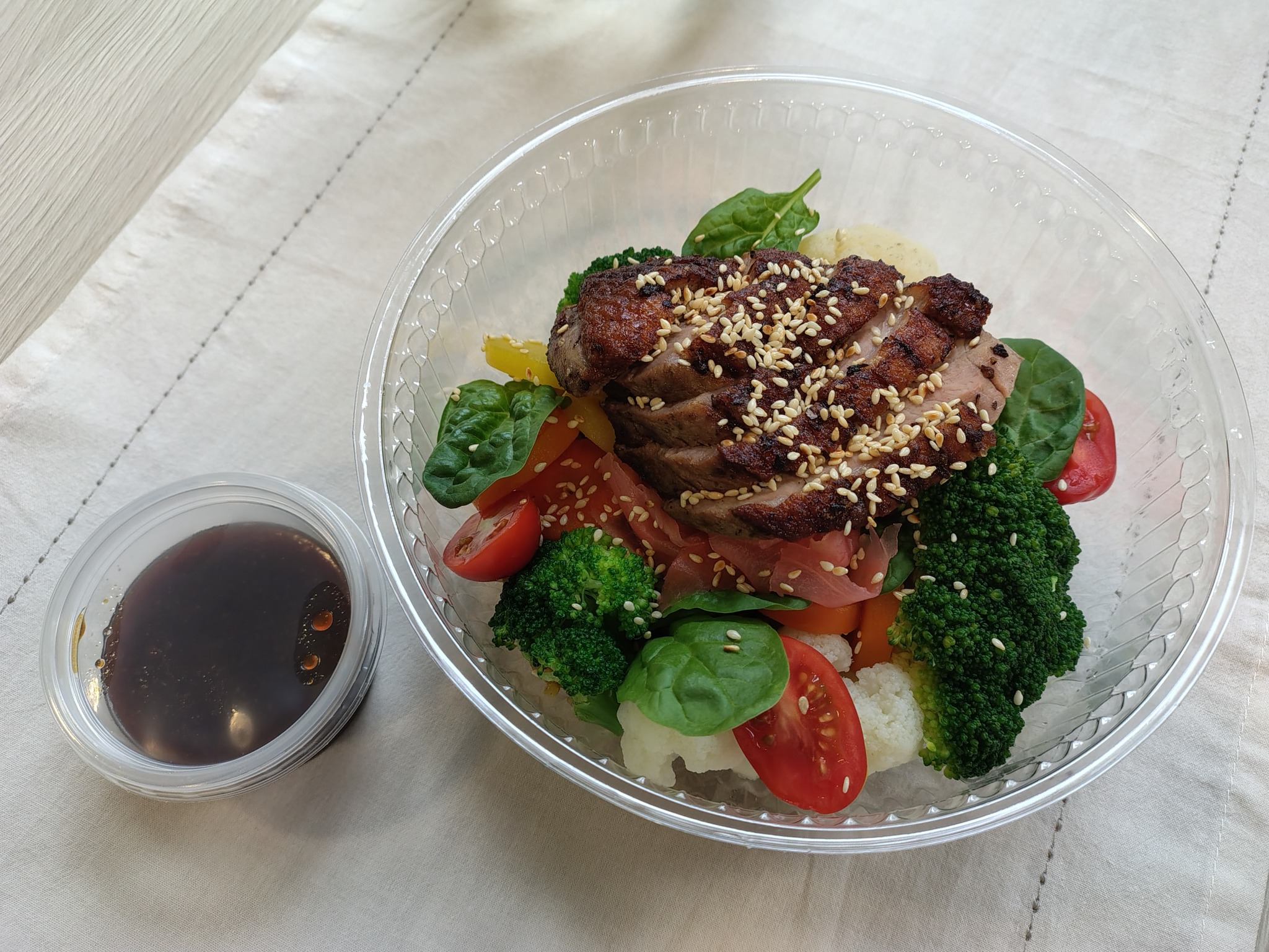 Gotowa sałatka z grillowaną kaczką posypaną sezamem, brokułami, kalafiorem, pomidorkami cherry, papryką i sosem w przezroczystym plastikowym pojemniku na białym obrusie.