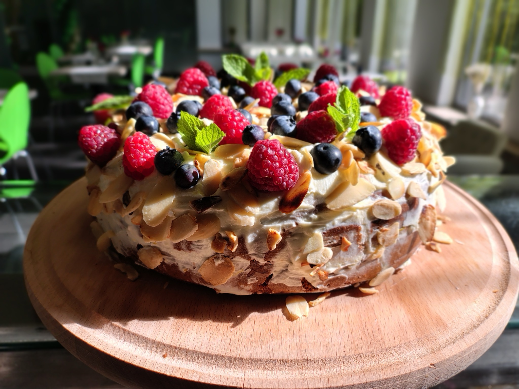 Tort z kremem, malinami, borówkami, migdałami i miętą na drewnianej tacy, w tle zielone krzesła.