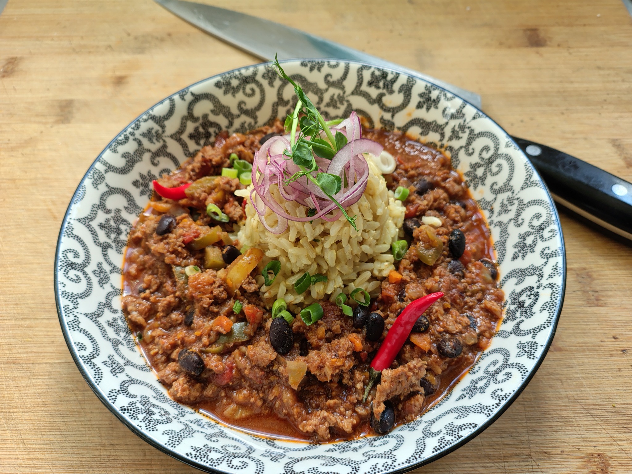 Chili con carne z ryżem, udekorowane czerwoną papryczką chili, zielonym szczypiorkiem i marynowaną czerwoną cebulą, podane na talerzu z czarno-białym wzorem na drewnianej desce. W tle widoczny nóż.