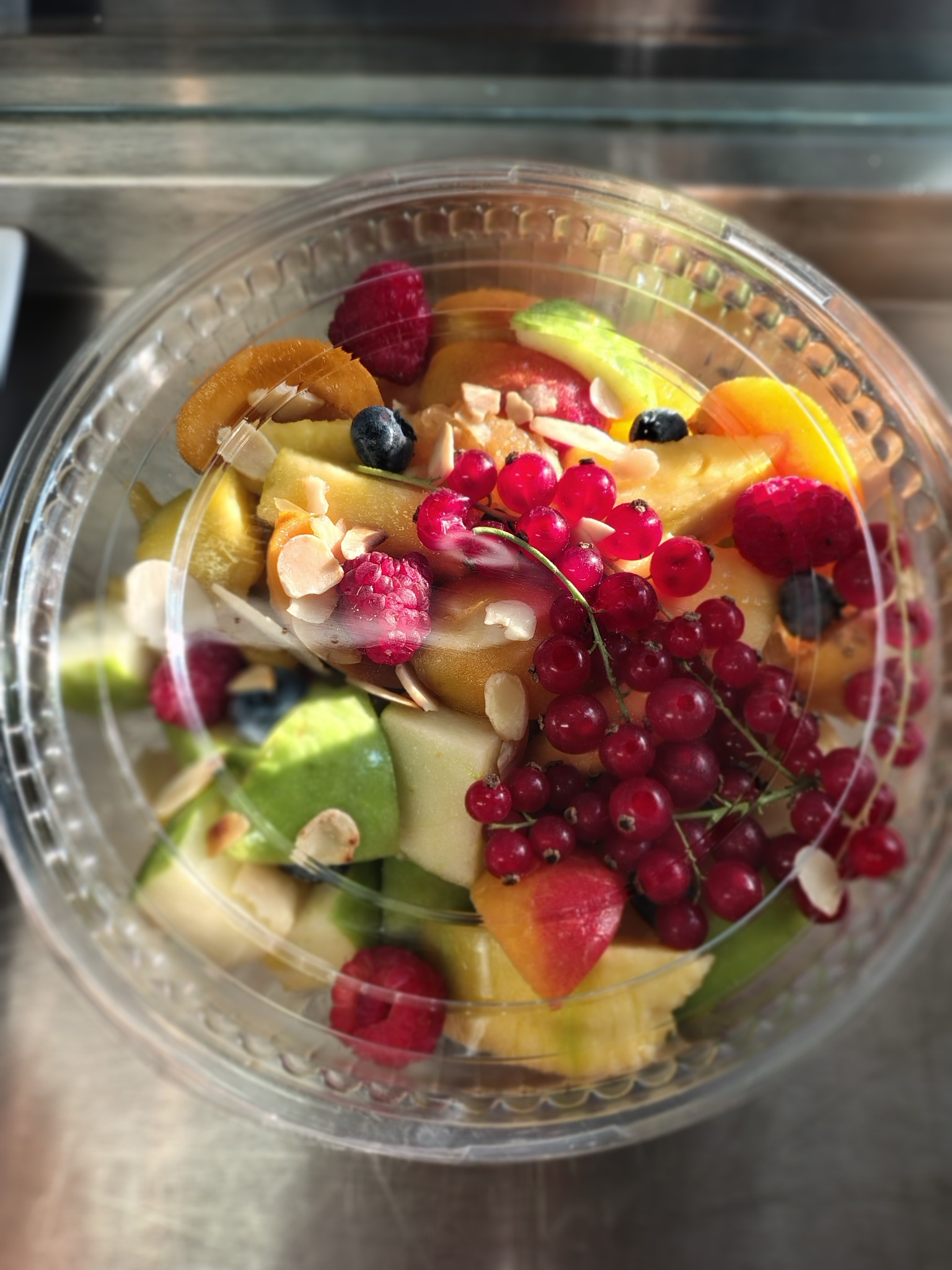 Widok z góry na sałatkę owocową w przezroczystym plastikowym pojemniku, zawierającą kawałki jabłek, ananasa, brzoskwiń, maliny, borówki i gałązki czerwonych porzeczek, posypaną płatkami migdałów.