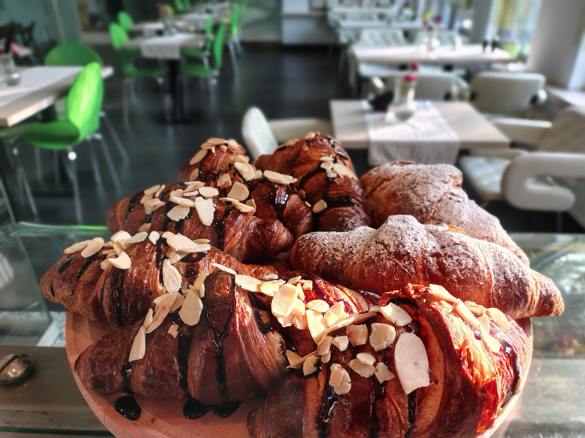 Kruche croissanty z czekoladą i płatkami migdałów, niektóre posypane cukrem pudrem, ułożone na drewnianej desce, w tle stoliki kawiarniane z zielonymi krzesłami.