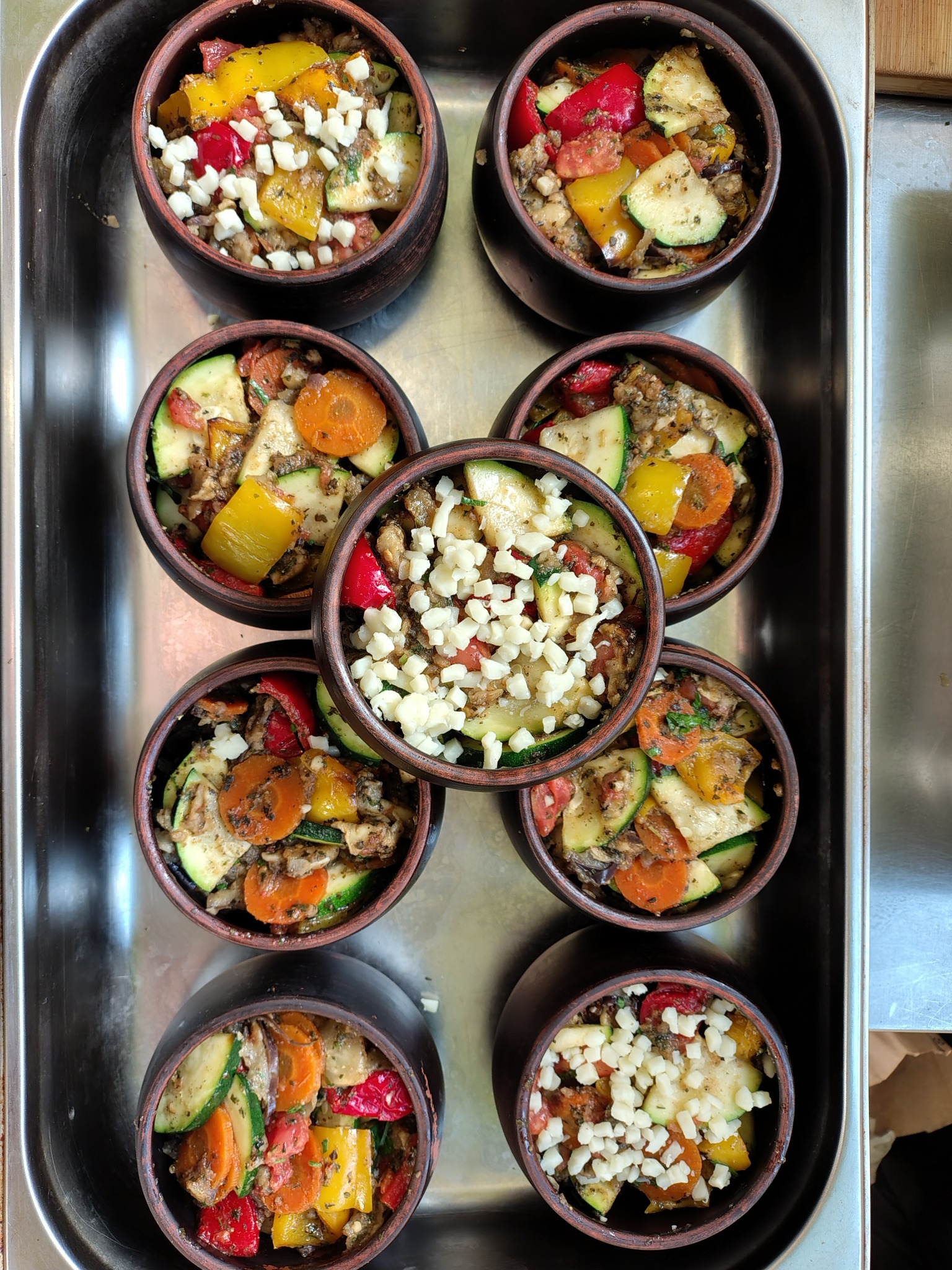 Glazurowane, pieczone warzywa (cukinia, marchew, papryka) w glinianych miseczkach na metalowej tacy; niektóre porcje posypane białym serem.