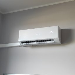 KLIREPO - Biały klimatyzator Haier zamontowany na szarej ścianie w pomieszczeniu, widoczna rura odprowadzająca skropliny w białej maskownicy.