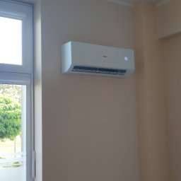 KLIREPO - Biały klimatyzator marki Haier zamontowany na beżowej ścianie w biurze, ustawiony na temperaturę 26 stopni Celsjusza, widoczne okno i lampa sufitowa.