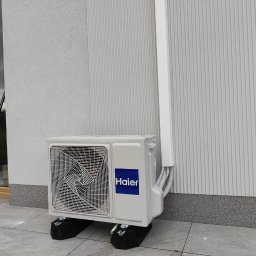KLIREPO - Zewnętrzna jednostka klimatyzacji Haier umieszczona na czarnych podkładkach antywibracyjnych, z białymi rurami odprowadzającymi skropliny przymocowanymi do elewacji budynku z oświetleniem zewnętrznym.