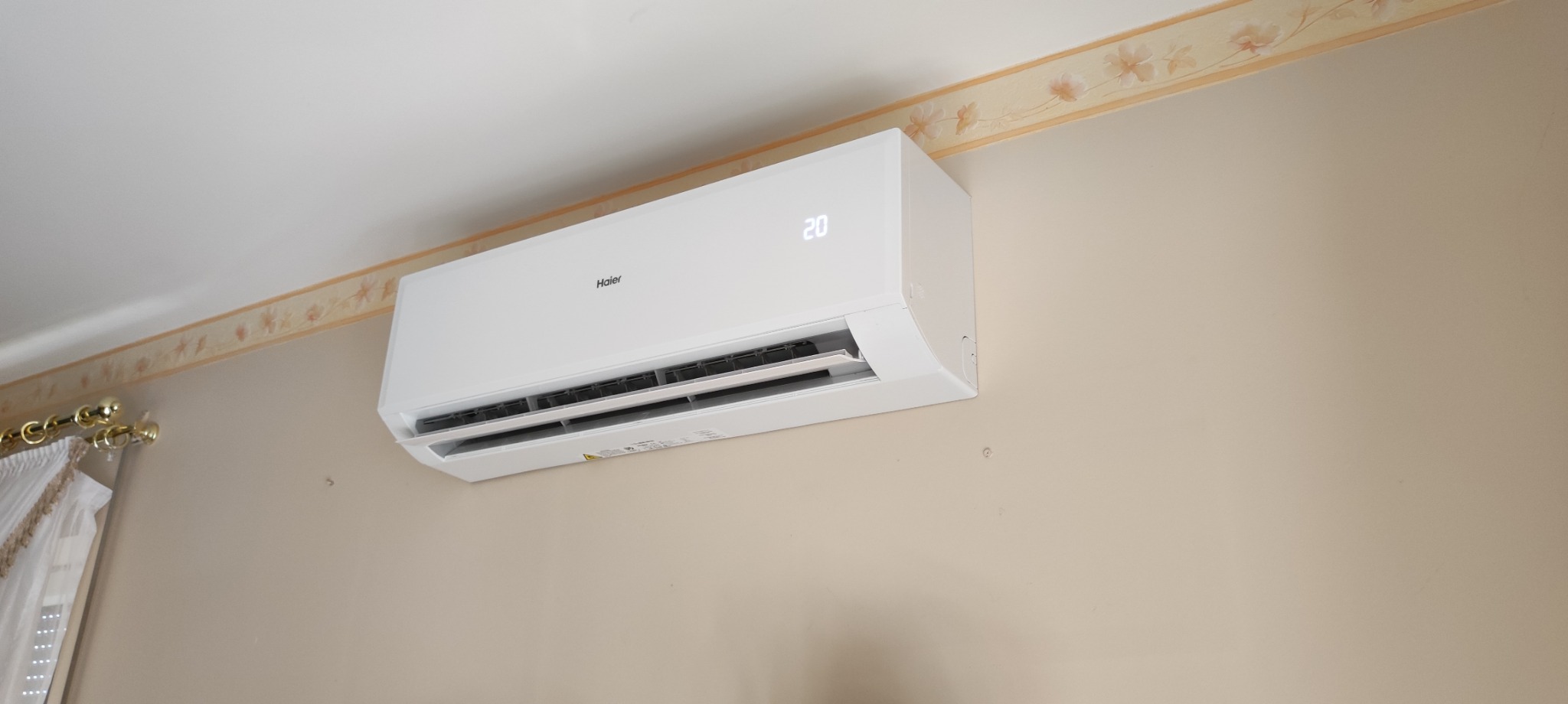 Biała jednostka klimatyzacji Haier zamontowana na beżowej ścianie z dekoracyjnym paskiem u góry, wyświetlająca temperaturę 20 stopni Celsjusza.