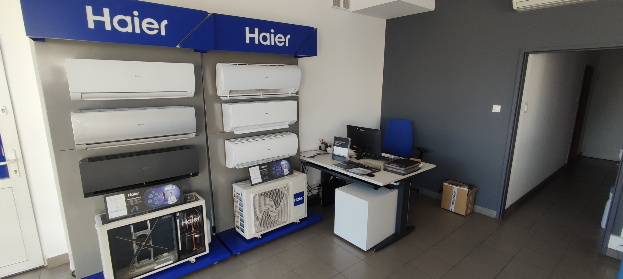 Ekspozycja klimatyzatorów Haier w salonie: modele ścienne w kolorze białym i czarnym, jednostka wewnętrzna z widocznymi elementami konstrukcyjnymi, biurko z komputerem i krzesłem w tle.