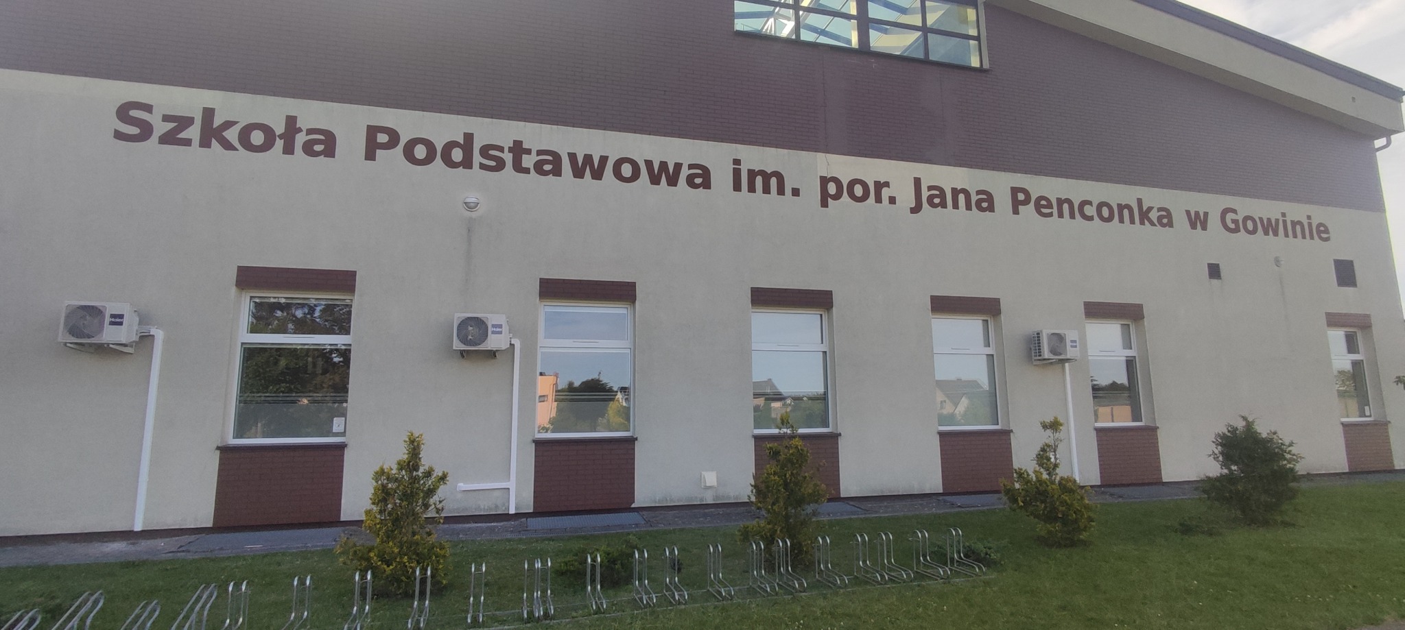 Zewnętrzne jednostki klimatyzacji zamontowane na ścianie budynku Szkoły Podstawowej w Gowinie, widoczne okna i stojaki na rowery.