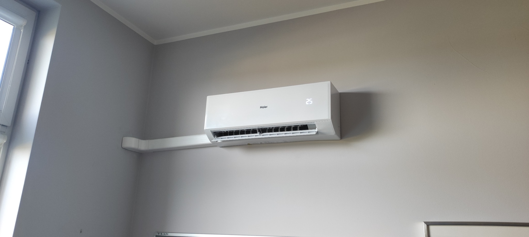 Biały klimatyzator Haier zamontowany na szarej ścianie w pomieszczeniu, widoczna rura odprowadzająca skropliny w białej maskownicy.