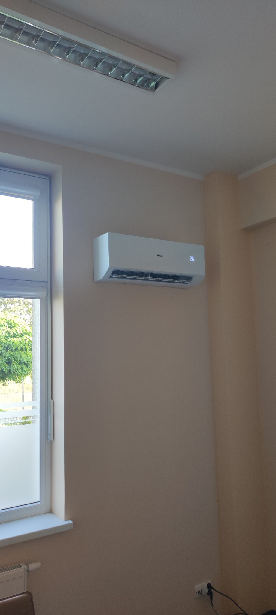 Biały klimatyzator marki Haier zamontowany na beżowej ścianie w biurze, ustawiony na temperaturę 26 stopni Celsjusza, widoczne okno i lampa sufitowa.