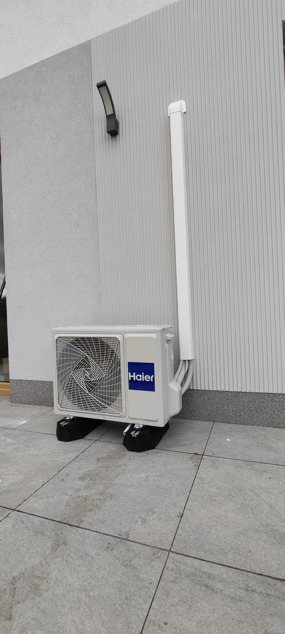 Zewnętrzna jednostka klimatyzacji Haier umieszczona na czarnych podkładkach antywibracyjnych, z białymi rurami odprowadzającymi skropliny przymocowanymi do elewacji budynku z oświetleniem zewnętrznym.
