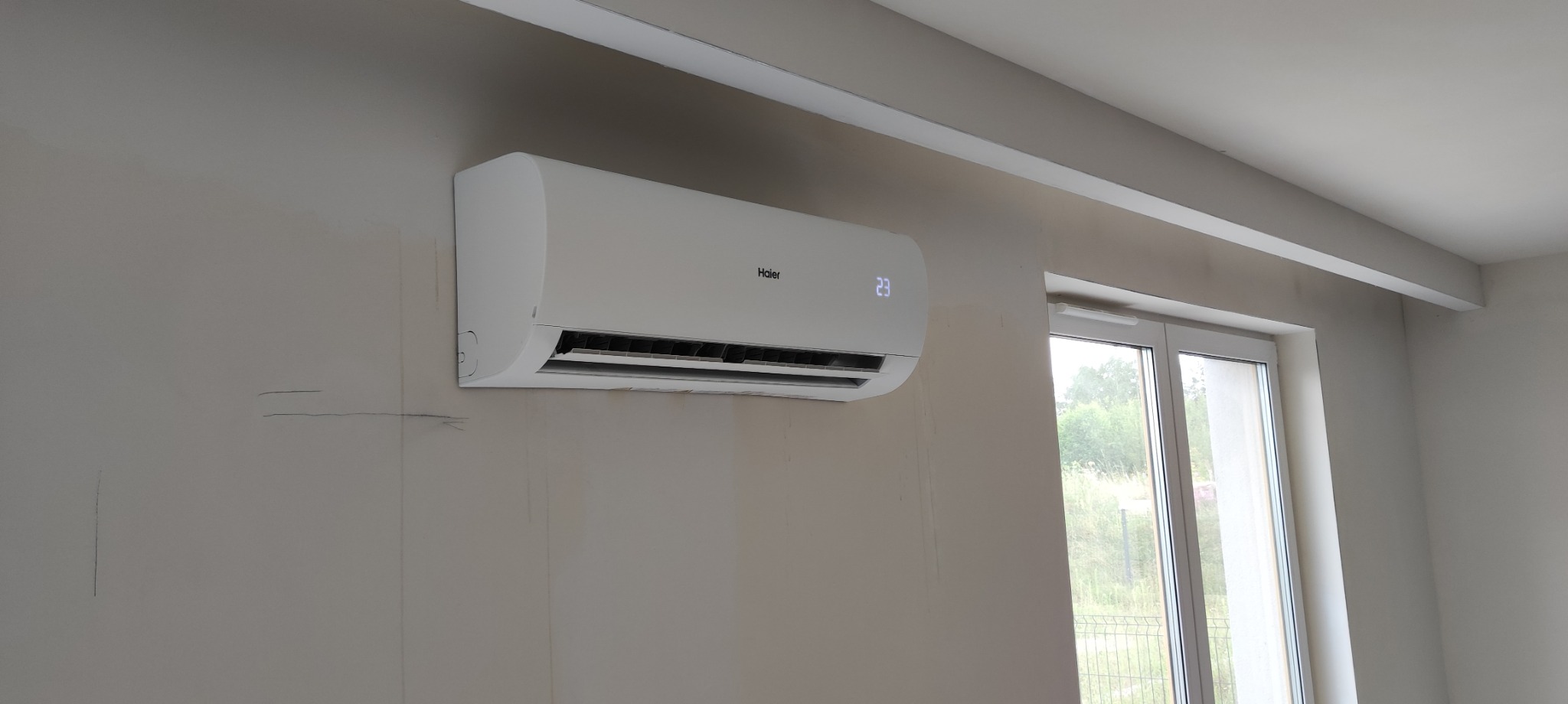 Biały klimatyzator Haier zamontowany na jasnoszarej ścianie w pomieszczeniu z oknem, widoczne ślady po montażu na ścianie.