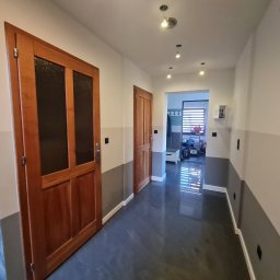 Tom-Bud - Wykończony korytarz z dwiema drewnianymi drzwiami z przeszkleniami, szarą lamperią na ścianach i ciemną, błyszczącą podłogą z płytek, widok na dalsze pomieszczenie z roletami w oknie.