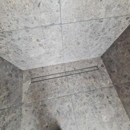 Tom-Bud - Szare płytki imitujące marmur na podłodze i ścianie, widoczny odpływ liniowy i fragment buta w dolnym rogu.