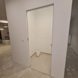 Tom-Bud - Wnętrze po remoncie z widocznymi świeżo wygładzonymi ścianami w odcieniach bieli, otwarte drzwi do małego pomieszczenia z workiem na podłodze, widoczny fragment podłogi przed i za drzwiami.
