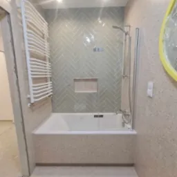 Wykończona łazienka z wanną obudowaną płytkami imitującymi lastryko, jasnoszare płytki ułożone w jodełkę na ścianie, biały grzejnik drabinkowy i owalne lustro w żółtej ramie, częściowo...