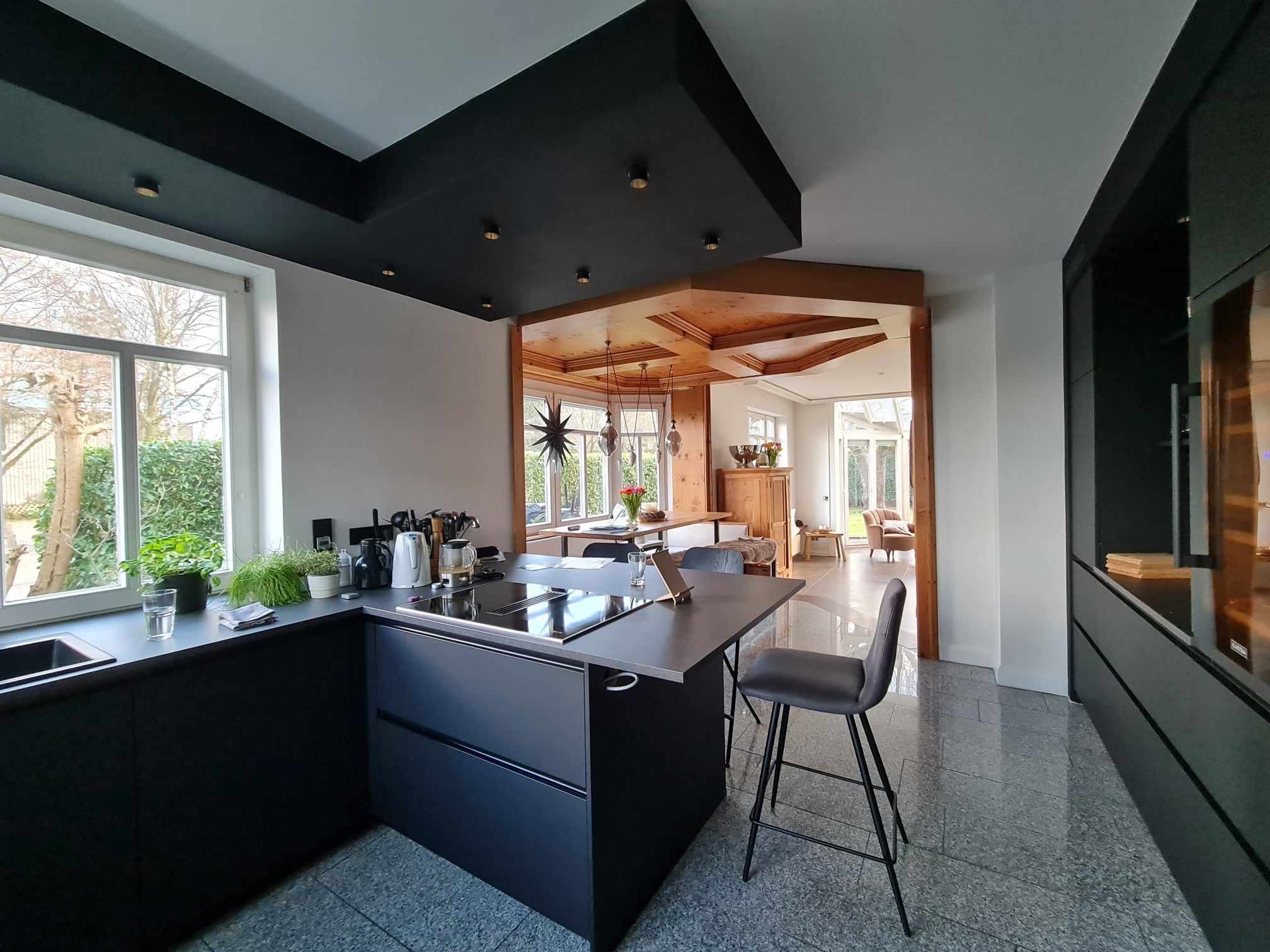 Nowoczesna kuchnia z czarną zabudową i wyspą kuchenną, szary blat, widok na jadalnię z drewnianym wykończeniem sufitu i jasne pomieszczenie w tle.