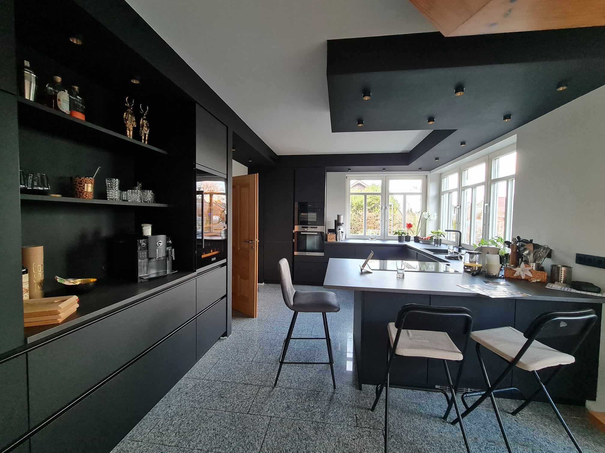 Przestronna, nowoczesna kuchnia w ciemnych kolorach z wyspą i oknami, oświetlona punktowo, z dekoracjami na półkach.